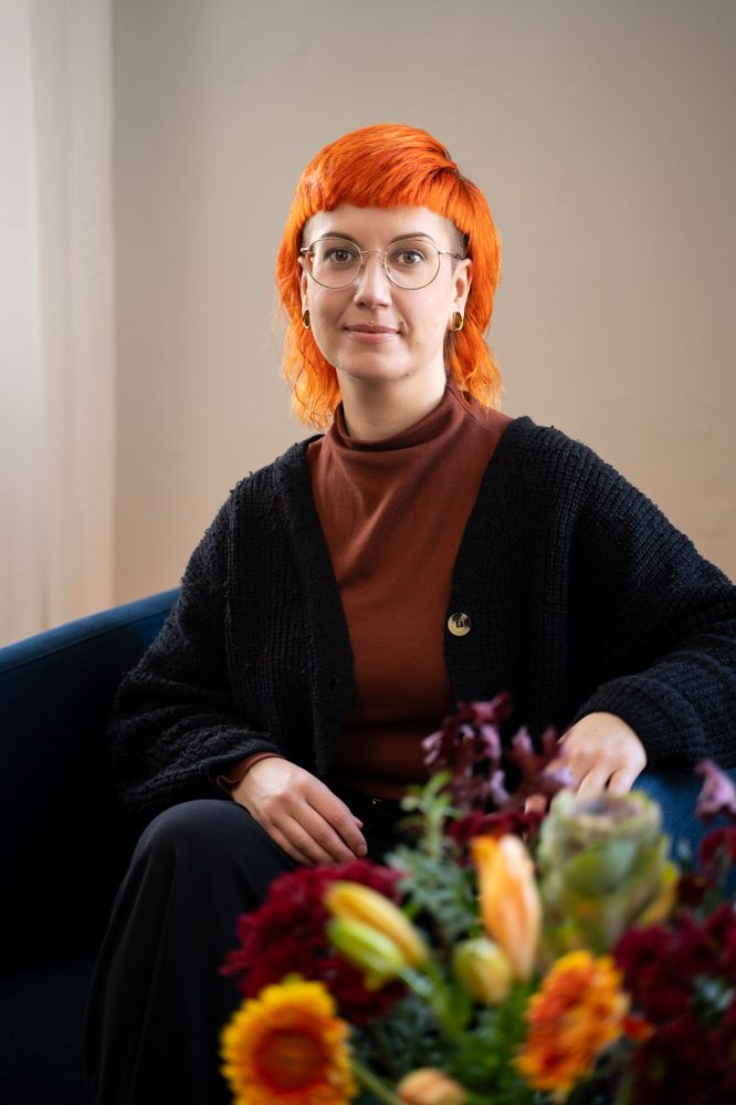 Willkommen Person mit orangefarbenem Haar, bräunlichem Pulli und Blumenarrangement auf einem Tisch schaut freundlich in die Kamera. Das ist Jule