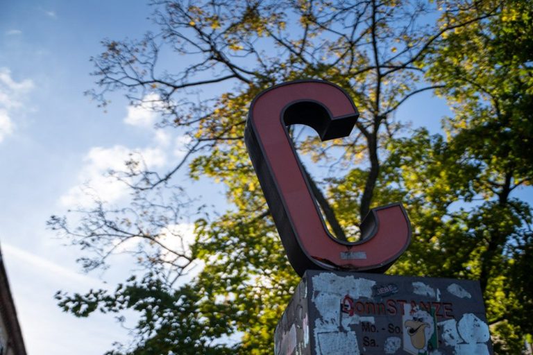 Neubeginn Roter, großer Buchstabe "C" vor blauem Himmel und grünen Ästen. Das ist der Hofeingang zur Praxis.